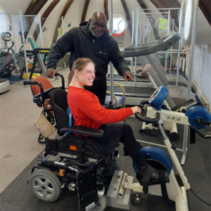 A power chair user smiles while using exercise equipment in the Ability Bow gym. A member of the Ability Bow team stands behind them.