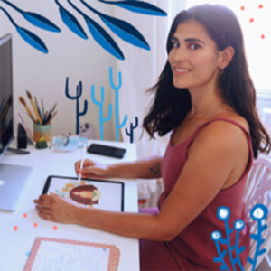 Ananya smiles at the camera while sitting at a desk. She is creating a digital illustration on a tablet. Illustrations of leaves and flowers have been added to the image.