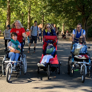 Skaters push children and adults in adapted wheelchairs in a sunny park.