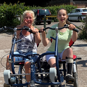 Two people ride an accessible bicycle while smiling at the camera.