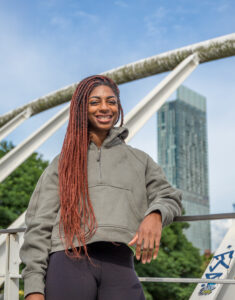 Kadeena Cox poses in front of a high rise building. Her hair is in long braids, and she wears a grey hoodie.