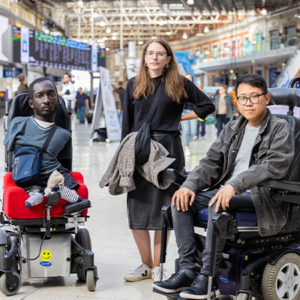 An image of three Transport for All campaigners standing in a busy train station. Two of the campaigners are wheelchair users.