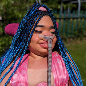Shelby smiles for the camera. She has blue and black braids in her hair, and wears a bright pink halterneck top.